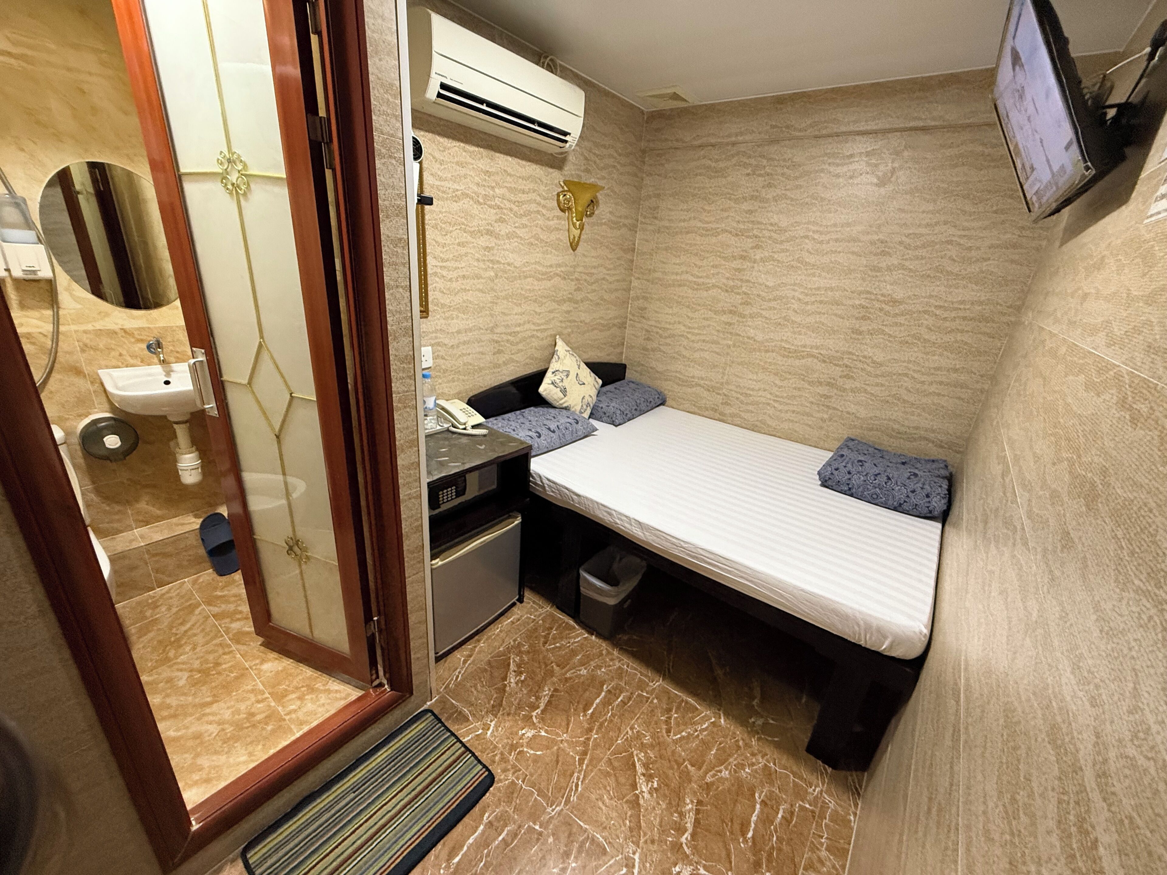 Habitación de hotel compacta con cama, un pequeño escritorio con teléfono, televisión instalada en la pared y baño con lavamanos e inodoro.