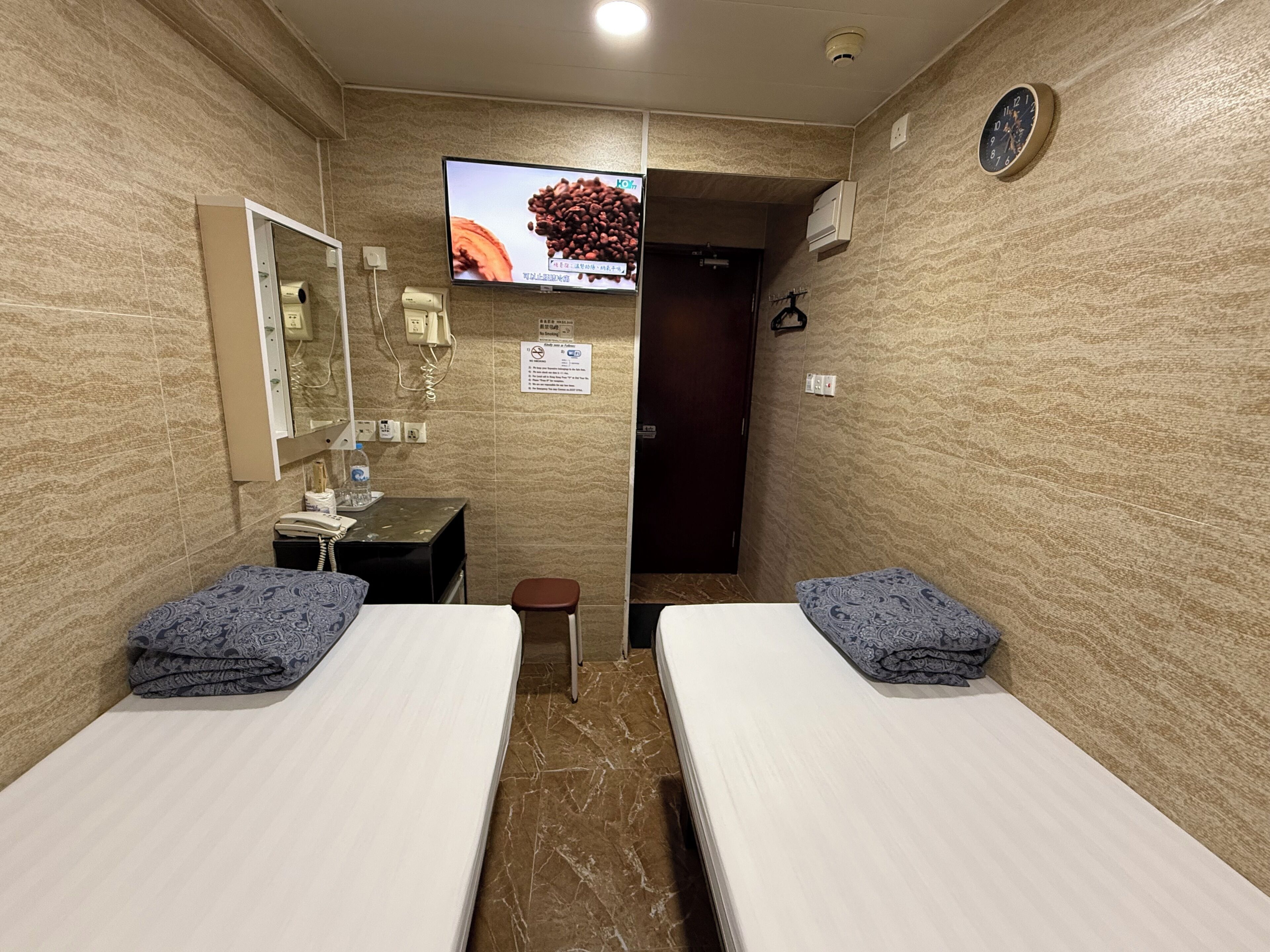 A hotel room with two beds, a small bathroom, a TV, and a clock on the wall.