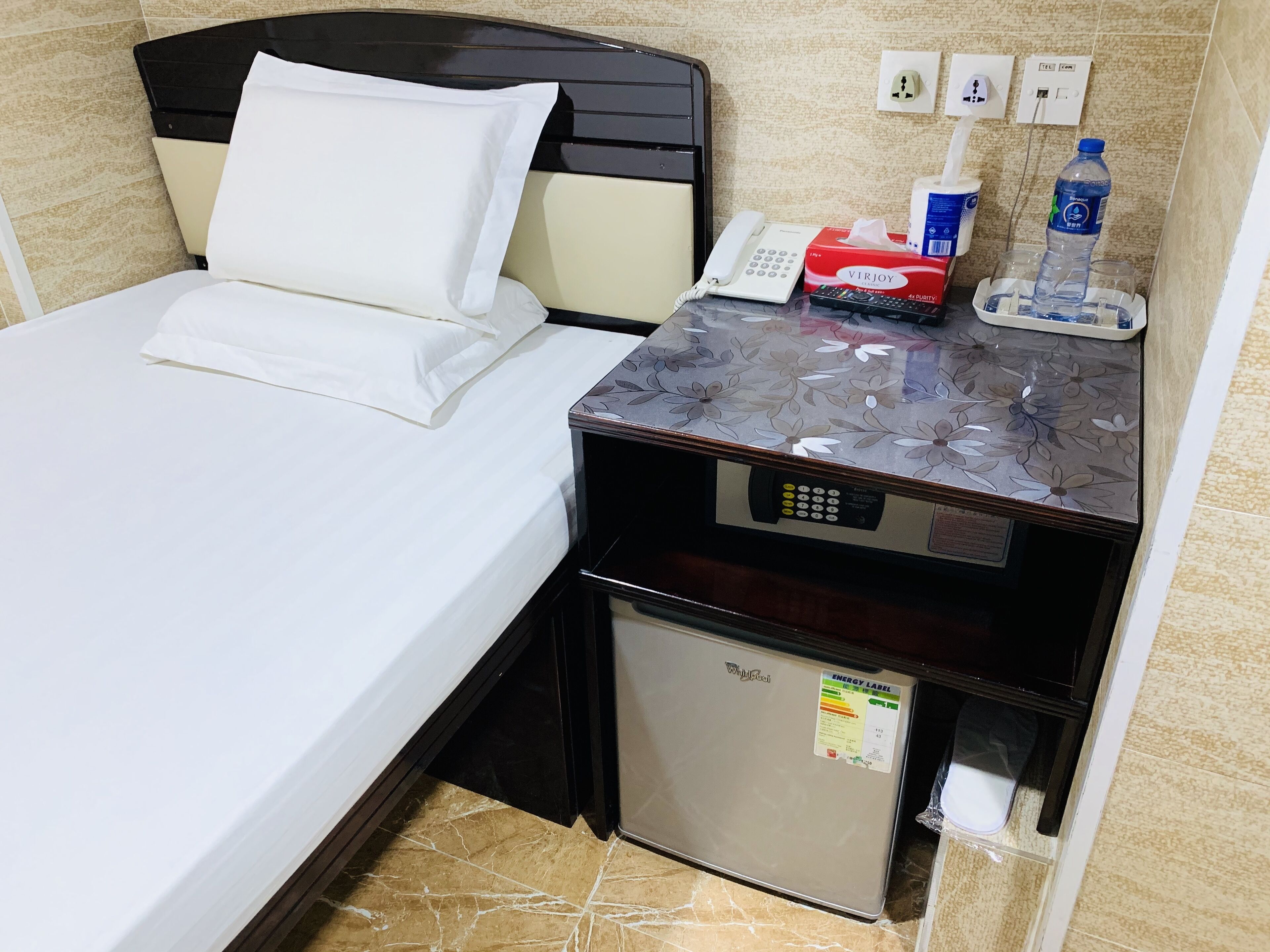 A hotel room with a bed, a bedside table with a phone, a tissue box, a water bottle, and a small refrigerator.