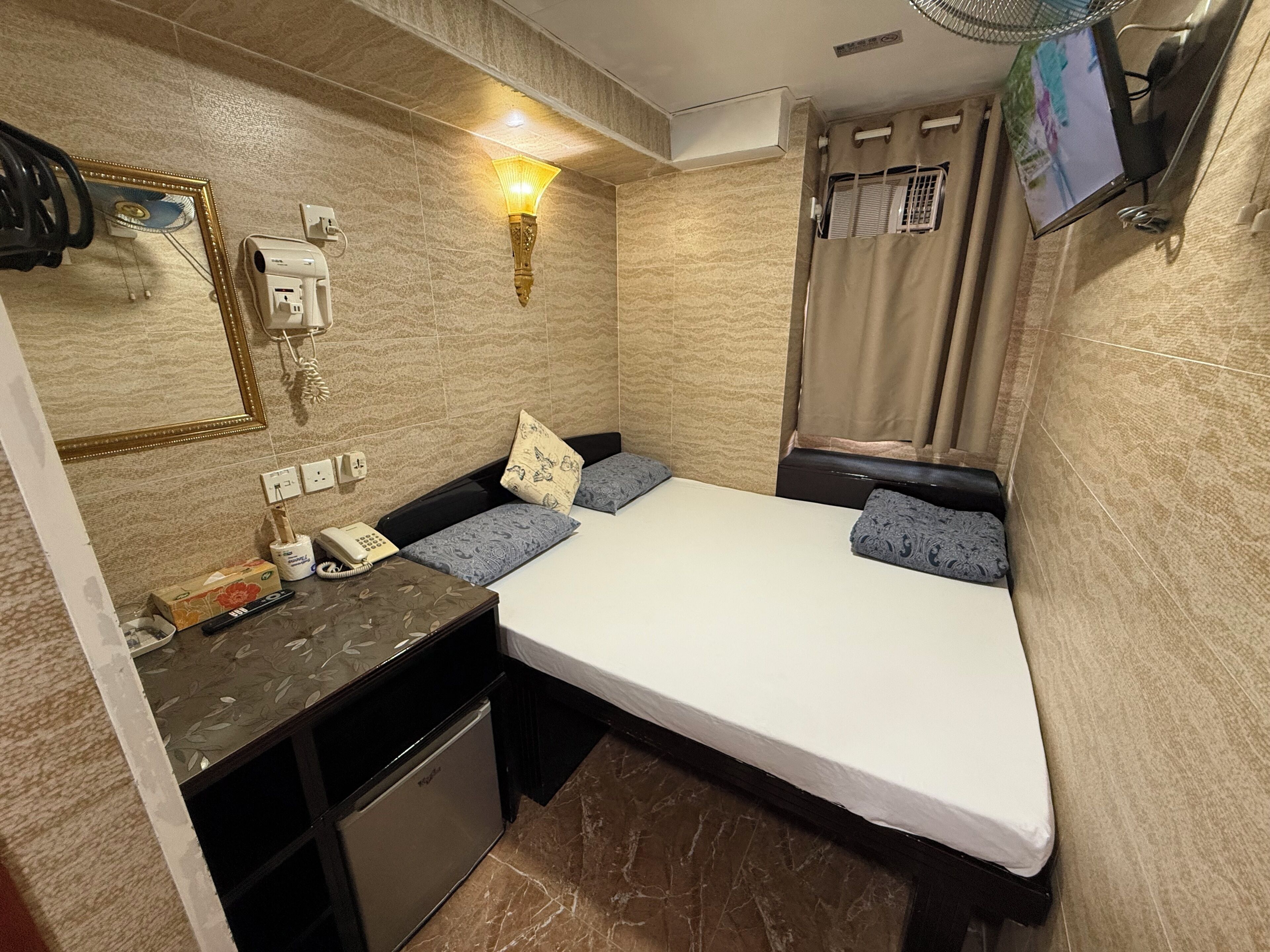 A compact hotel room with a bed, a small desk, a telephone, a fan, and a TV mounted on the wall.