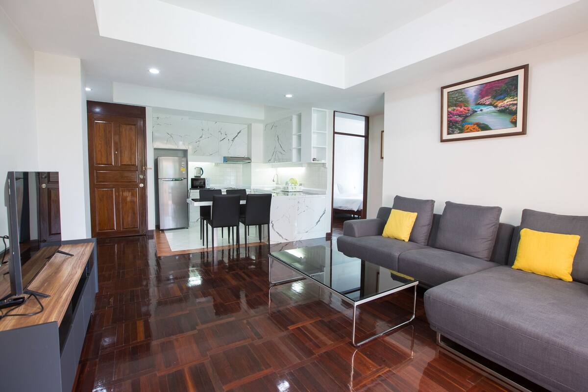 Ein modernes Wohnzimmer mit einem grauen Sofa, einem Glastisch und einem Holzbüro. Im Hintergrund befindet sich eine Küche mit weißen Schränken und ein Essbereich mit Stühlen.