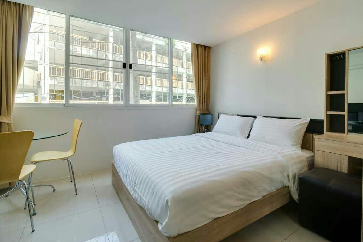 Ein Hotelzimmer mit Bett, Schreibtisch, Stuhl, Nachttisch, Lampe und einem großen Fenster mit Blick auf Gebäude.