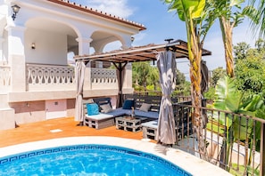 Un espace aménagé au bord de la piscine, comprenant un coin salon couvert, une piscine et une végétation luxuriante.