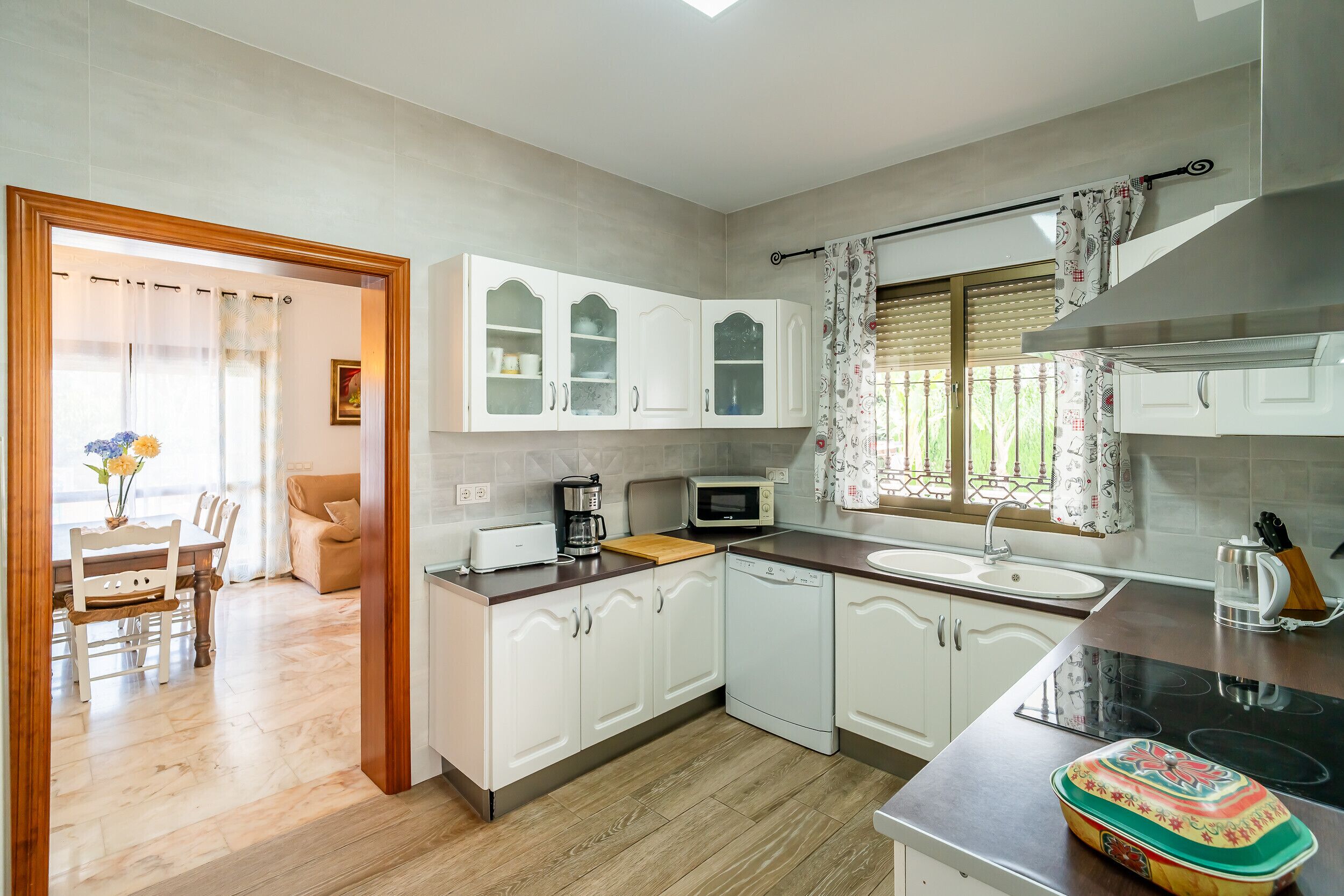 A modern kitchen with white cabinetry, a dining area with a wooden table and chairs, and a view of the outdoors through a window with patterned curtains.