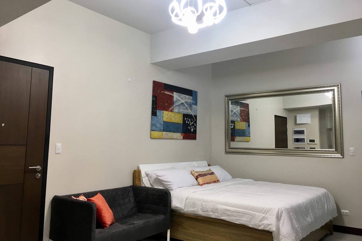 A bedroom with a bed, a black sofa, a colorful abstract painting, and a large mirror.