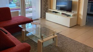 Een woonkamer met een rode bank, een glazen salontafel, een tv op een witte dressoir, en uitzicht op buiten door een schuifdeur.