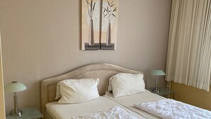 Een hotelkamer met een tweepersoonsbed, twee nachtkastjes met lampen, een beige vloerkleed en twee wanddecoraties van vazen.