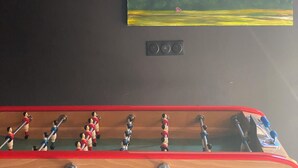 Un tableau représentant un paysage avec des nuages, un champ et des arbres, fixé au mur. En dessous du tableau, on trouve un panneau de commande avec trois boutons.