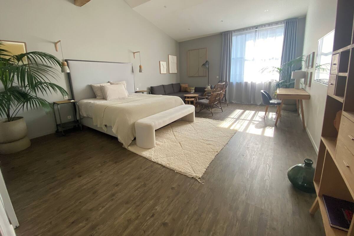 A bedroom with a large bed, a desk, a chair, a sofa, a lamp, and a potted plant.