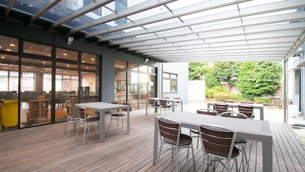 An outdoor patio area with tables and chairs, a glass ceiling, and a view of the building's interior.