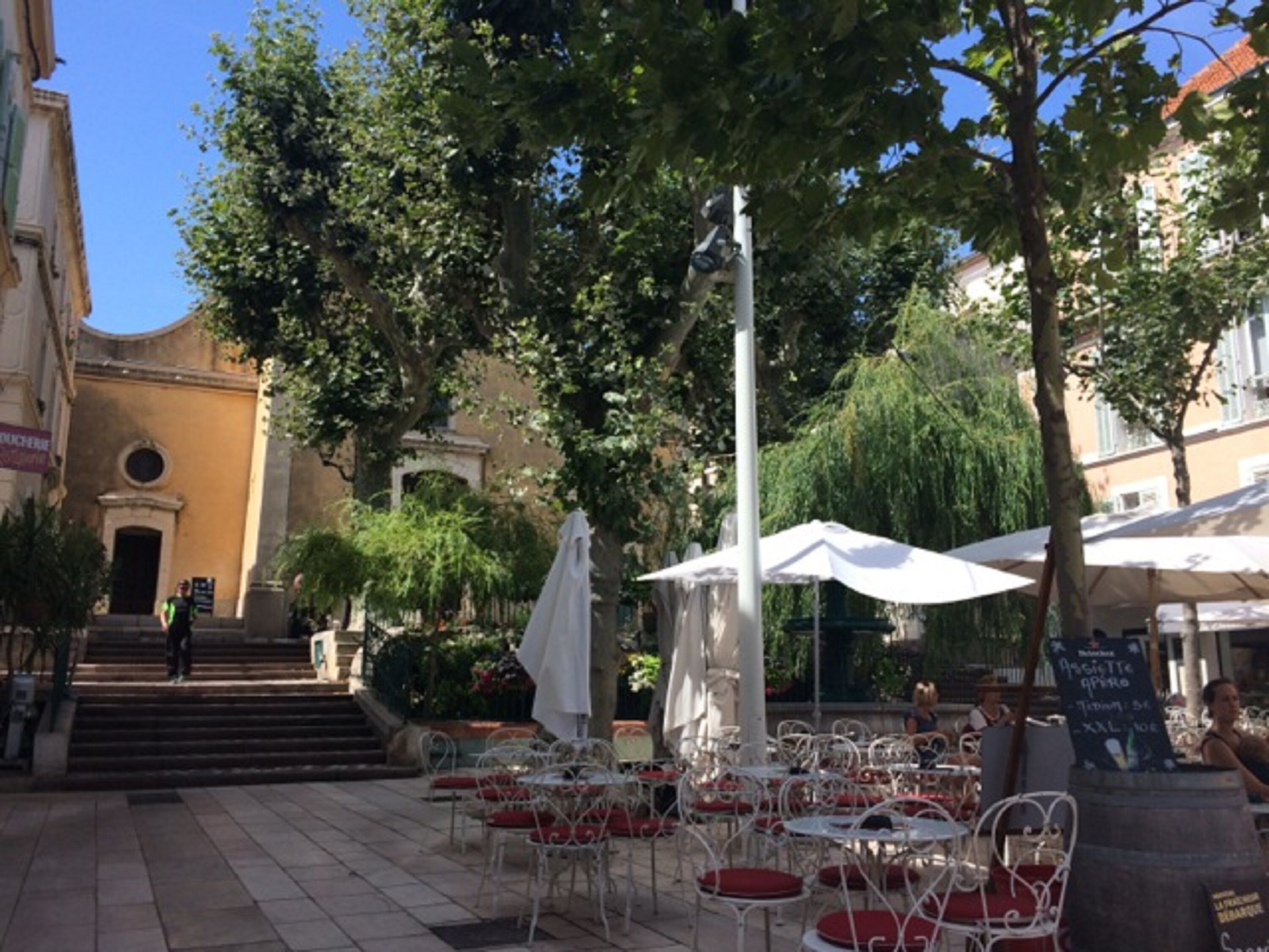 An outdoor café area with tables and chairs, surrounded by trees and buildings.
