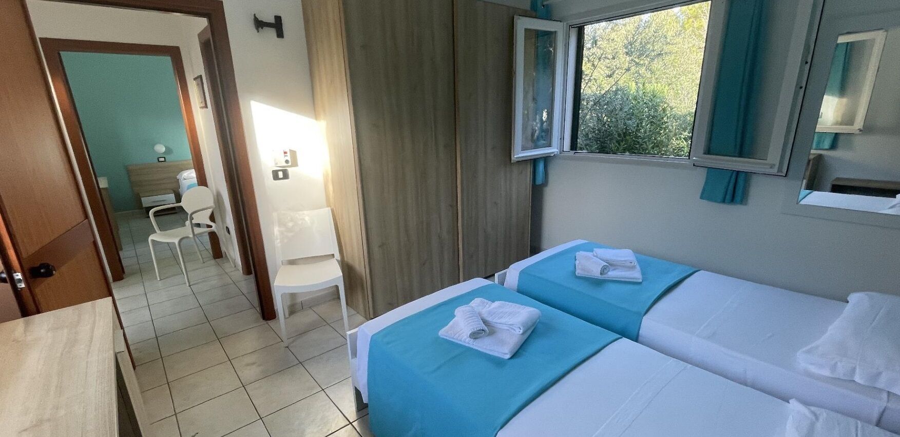 A hotel room with two beds, a small desk, a chair, and a window with curtains.