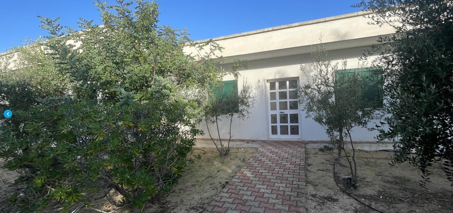 A building with a paved entrance, a window with shutters, and surrounding greenery.