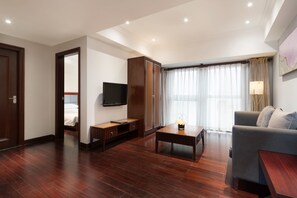 A modern hotel room with a wooden floor, a grey sofa, a TV, a wooden cabinet, and a large window with curtains.