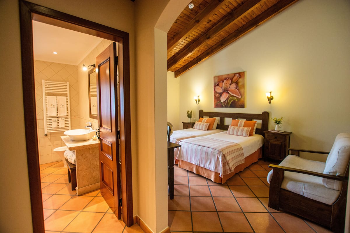A hotel room with a bathroom, a bed with pillows, a chair, and a painting on the wall.