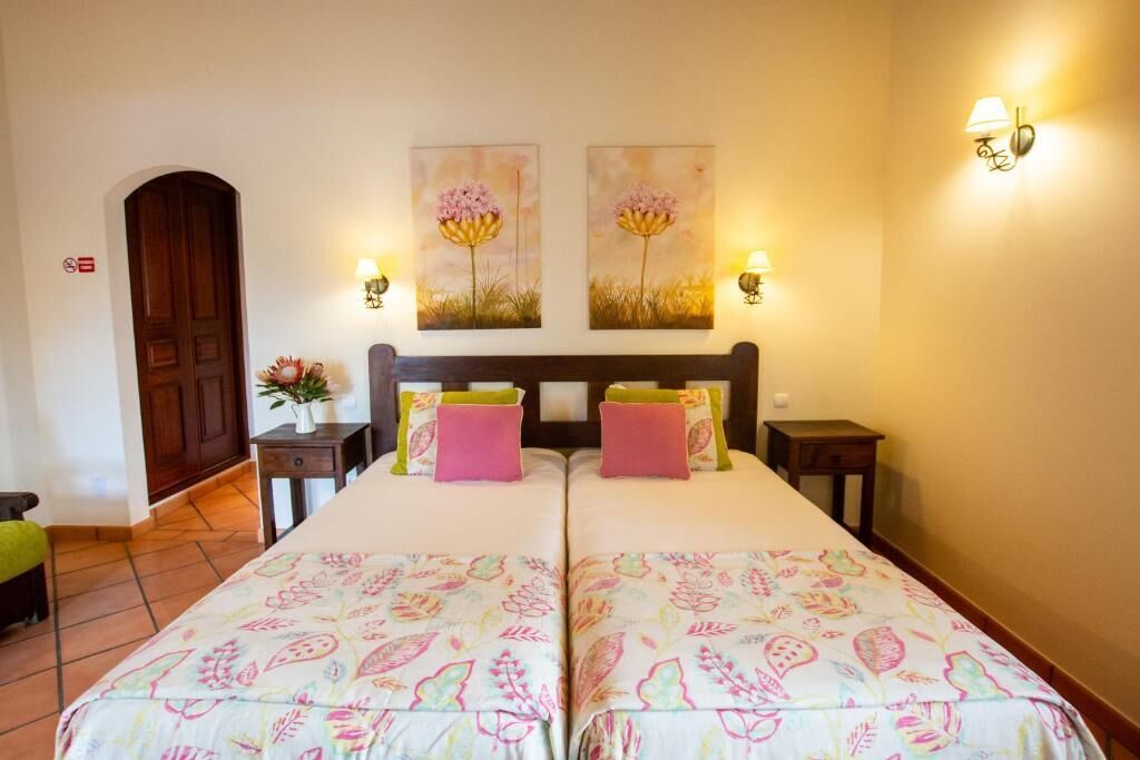 Een kamer met twee bedden, een houten deur, een kleine tafel met een bloevas en twee wandlampen.