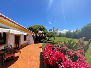 A house with a patio, a dining table, chairs, and a garden with pink flowers.