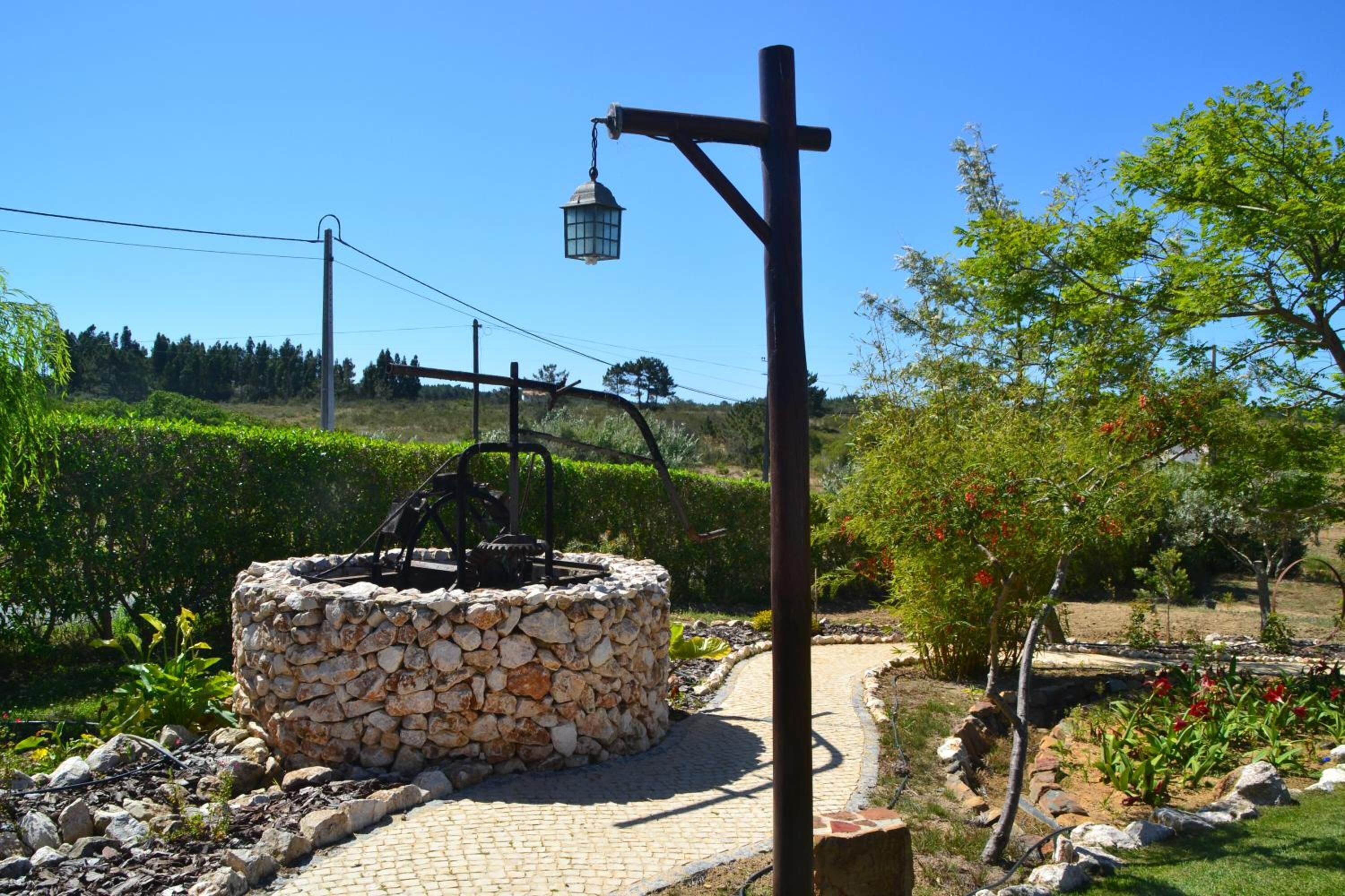 Un puits en pierre, surmonté d’une pompe en métal, entouré d’un chemin pavé et de verdure.