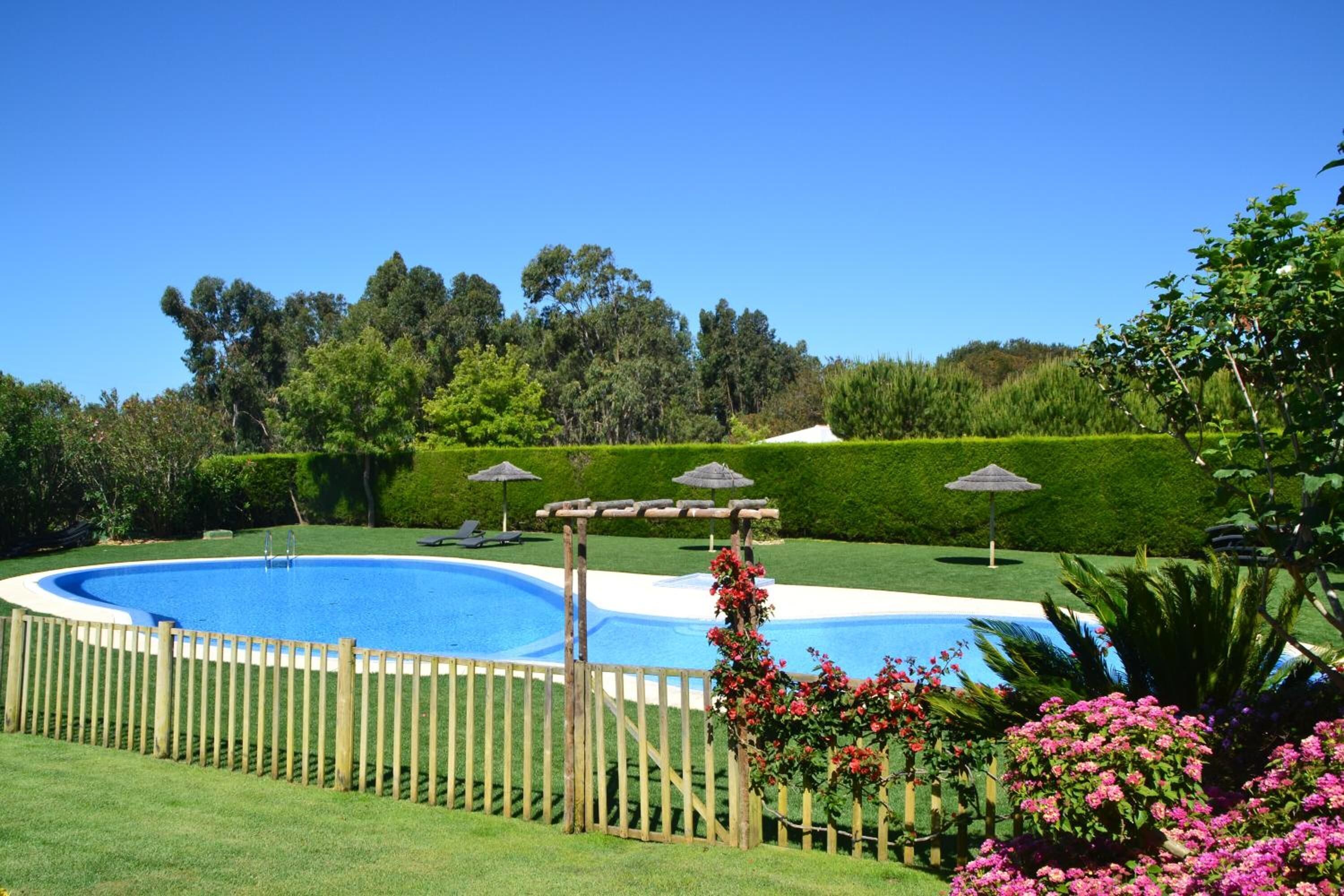 A swimming pool surrounded by a wooden fence, with a pergola and umbrellas in the background.