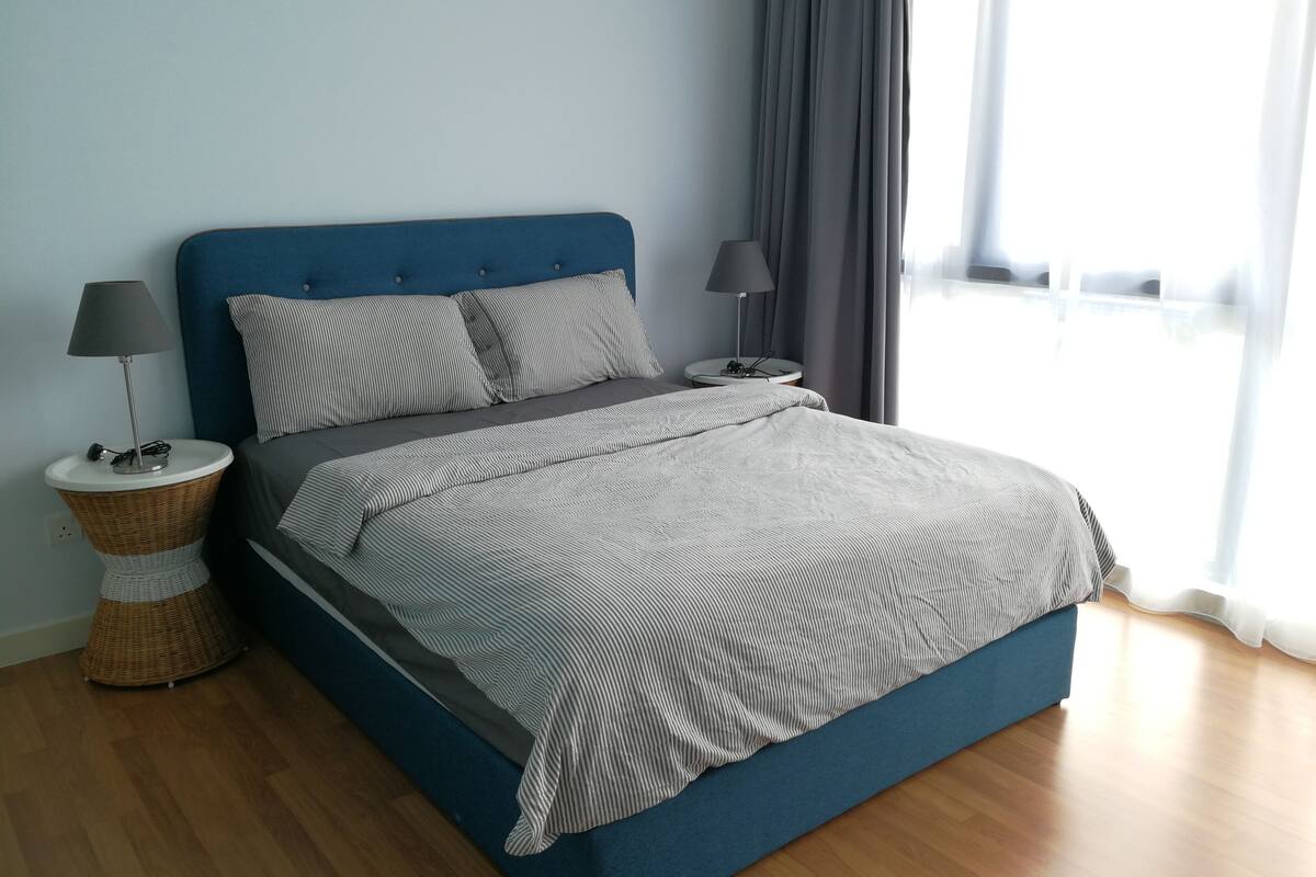 A modern bedroom with a blue headboard, two bedside tables with lamps, a wicker side table, and a large window with curtains.