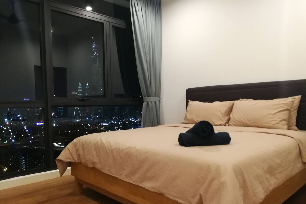 A bedroom with a large bed, a view of a cityscape through a window, and a neatly made bed with a rolled towel.
