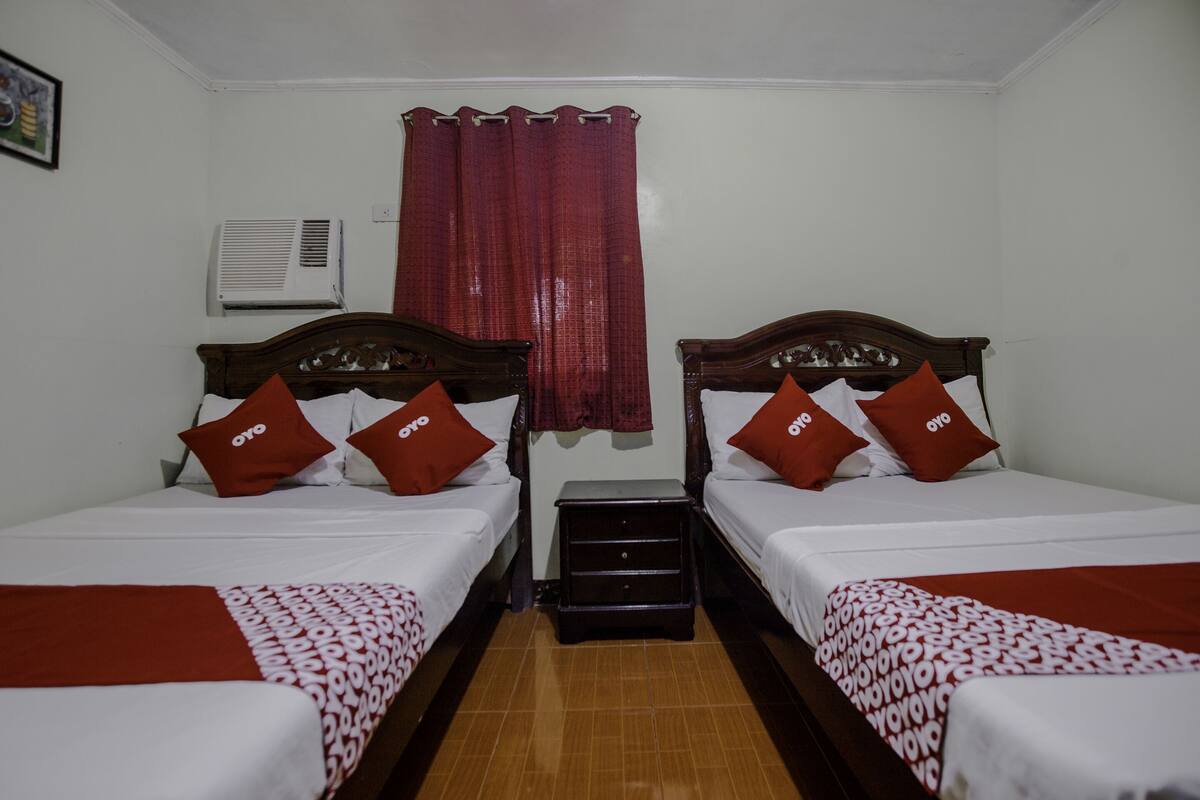 Habitación de hotel con dos camas individuales, cada una con cabecero de madera, ropa de cama blanca y almohadas rojas. Entre las camas hay una mesita de noche de madera, y se ve una cortina roja al fondo.