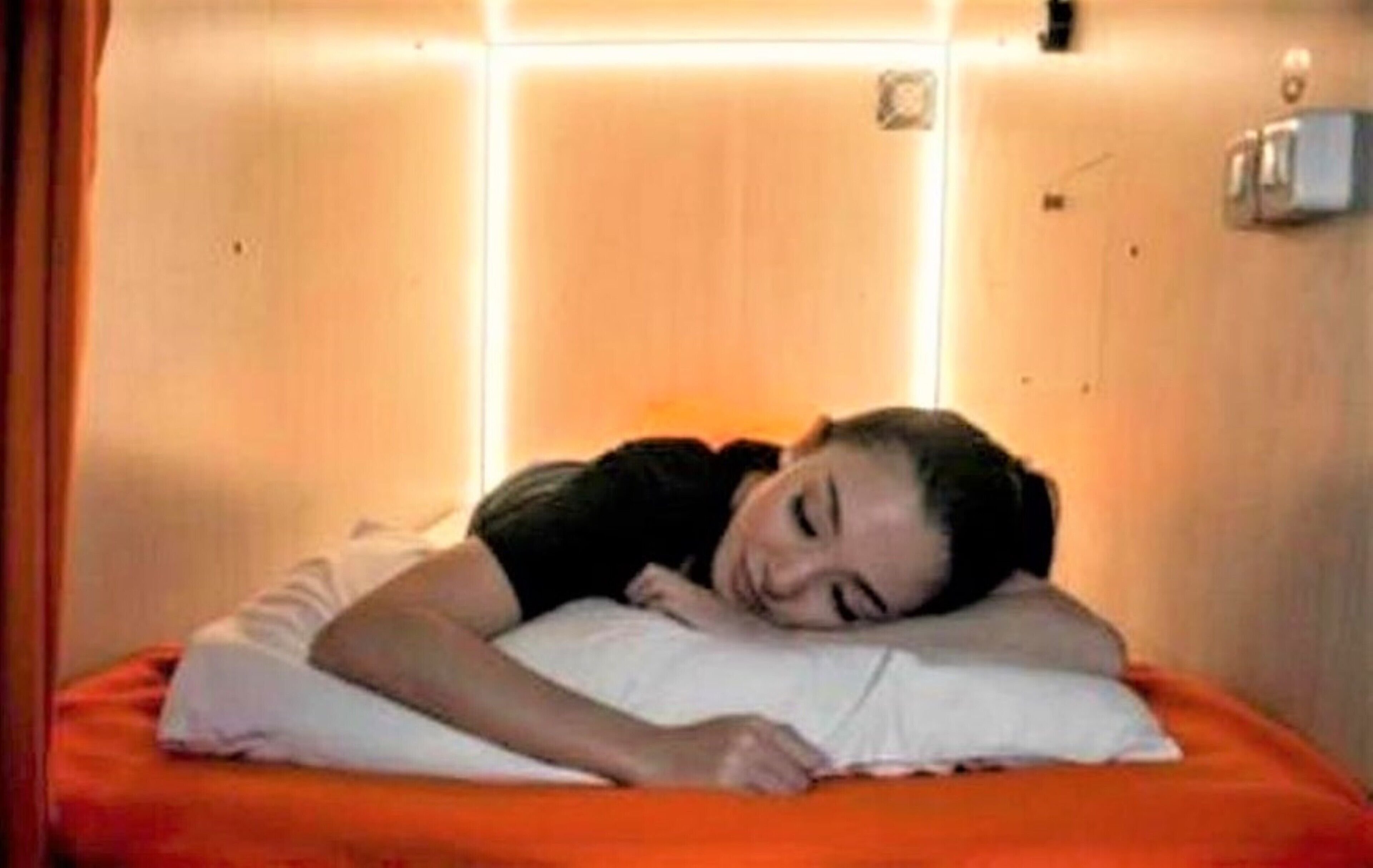 A person sleeping in a room with a bed, a wall-mounted light, and a thermostat.