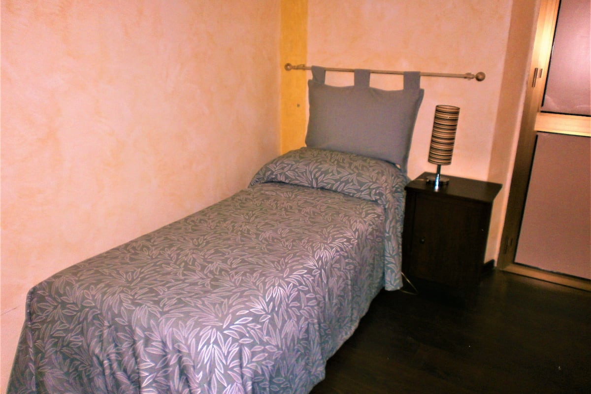 A single bed with a patterned bedspread, a bedside lamp, and a small nightstand in a room with plain walls.