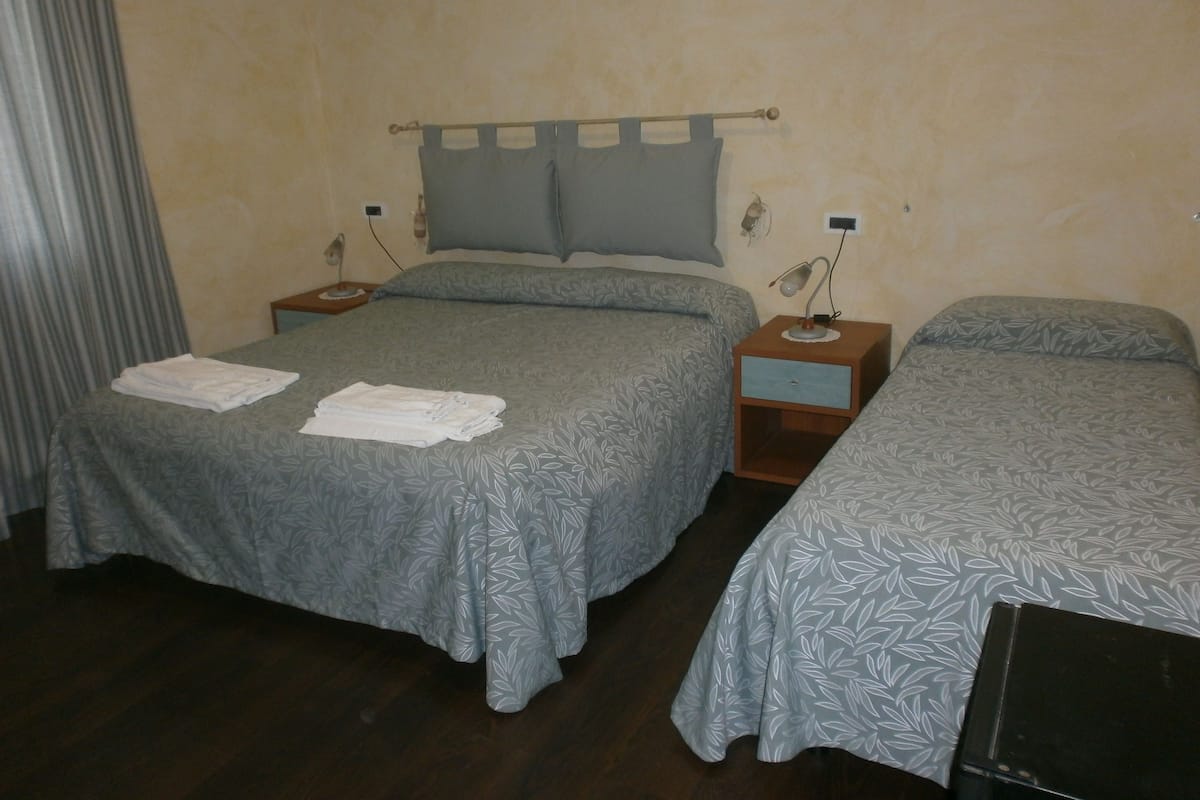 A bedroom with two beds, each with a grey floral bedspread, a wooden nightstand with a lamp, and a wall-mounted shelf.