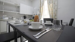 Una mesa de comedor con una taza blanca, plato, vaso y cubiertos, en una cocina moderna con armarios blancos y un ventanal con cortinas.
