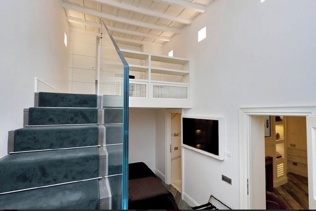 A modern interior with a glass staircase leading to a mezzanine, a flat-screen TV mounted on the wall, and a wooden floor.