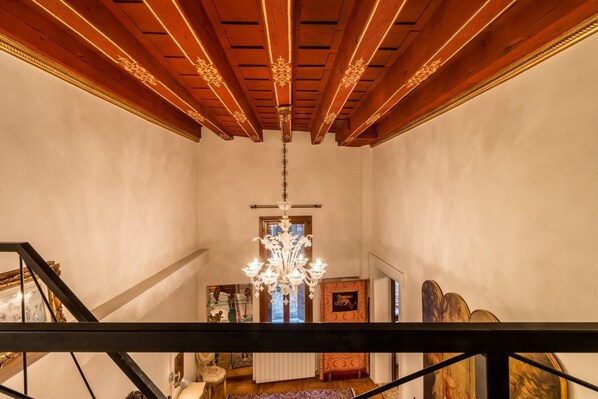 A grand staircase with a chandelier, wooden ceiling beams, and a view of a room with a patterned rug and artwork.