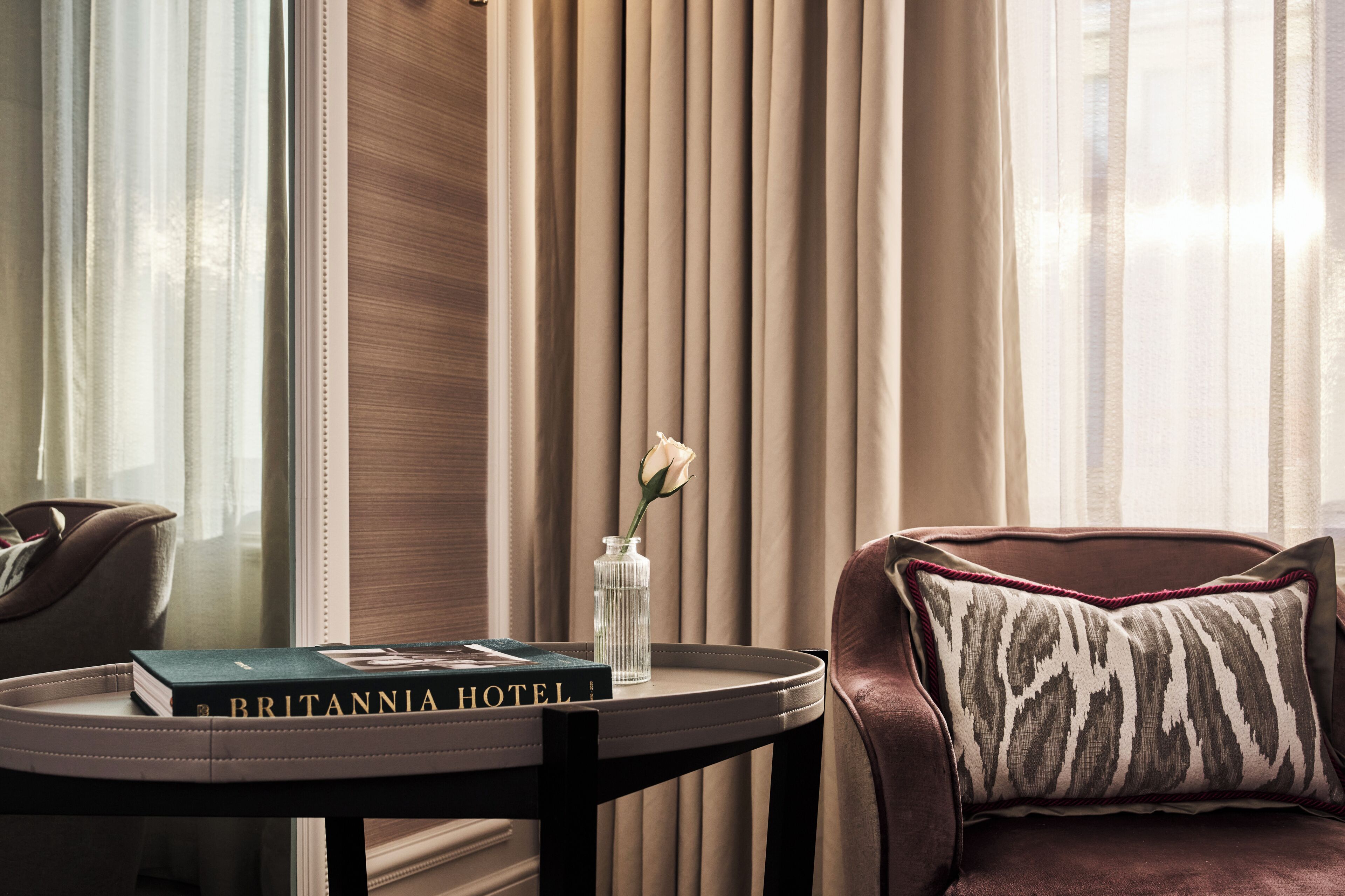 Un divano in pelle con un cuscino decorato, un tavolino con un libro intitolato 'BRITANNIA HOTEL' e un vaso con un singolo fiore bianco.