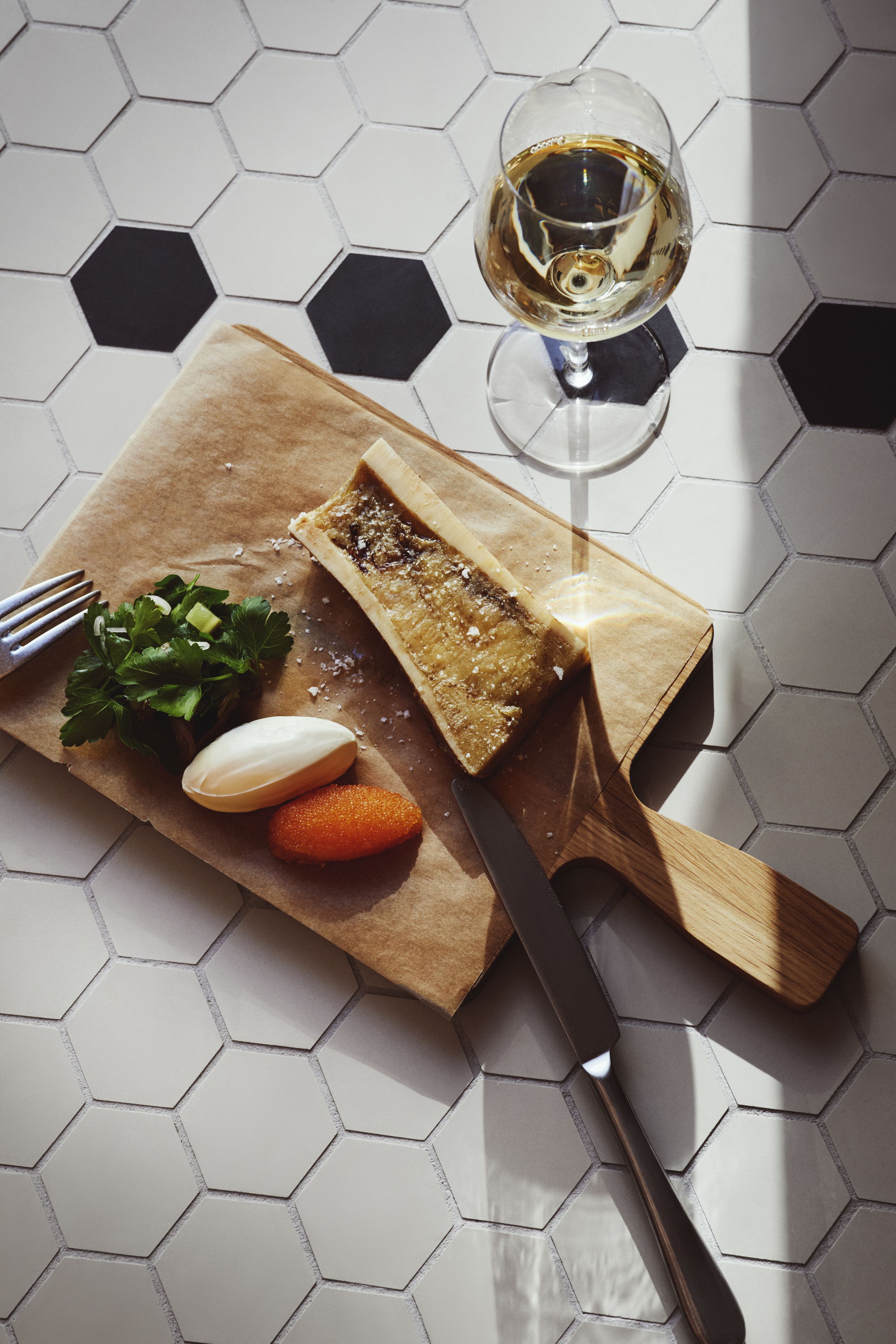 A wooden cutting board with a piece of bread, a knife, a glass of white wine, and some vegetables on a tiled floor.