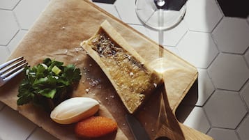 A wooden cutting board with a piece of bread, a knife, a glass of white wine, and some vegetables on a tiled floor.