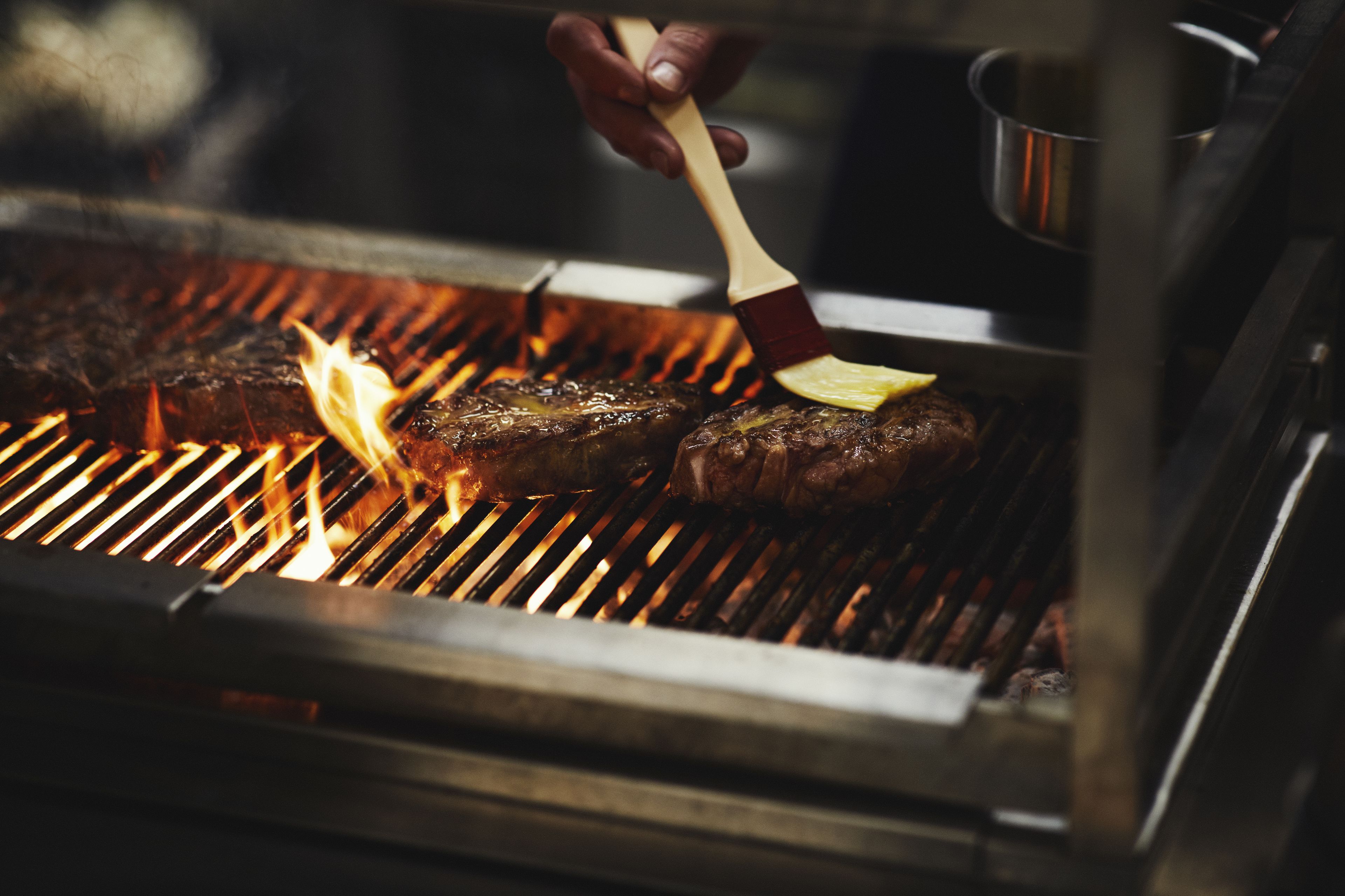 A close-up of a grill with meat being cooked, a hand holding a brush, and a pot in the background.