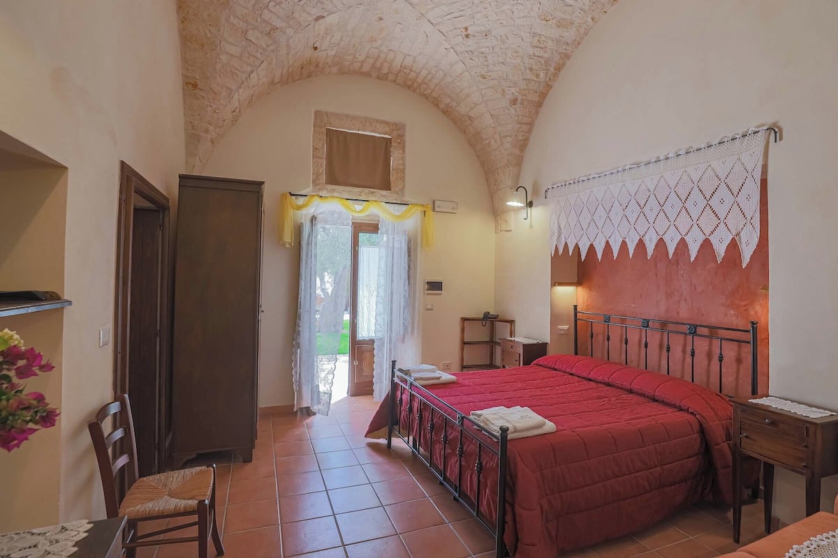 Une chambre à coucher avec un lit rouge, une chaise en bois, une petite table de chevet et une fenêtre avec des rideaux.
