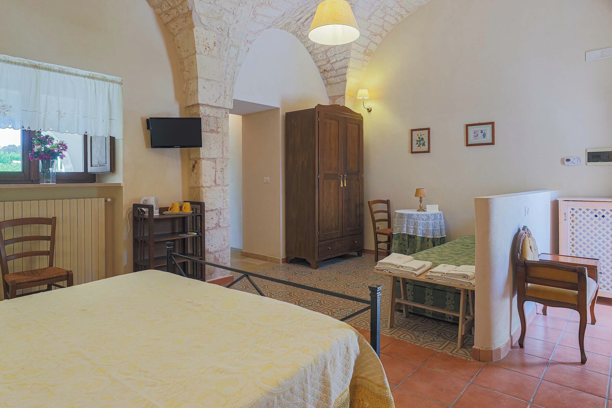 Una habitación con una cama, un armario de madera, una mesa con una silla, una mesita de noche con una lámpara y una ventana con cortina.