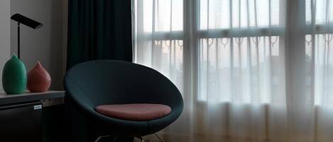A room with a modern chair, a desk, and a window with sheer curtains.