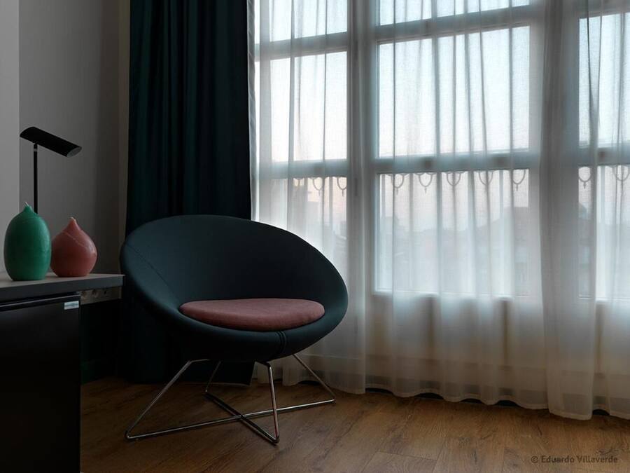 Una habitación con una silla moderna, un escritorio y una ventana con cortinas translúcidas.