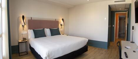 A hotel room with a large bed, bedside lamps, a small table with a chair, and a door leading to another room.