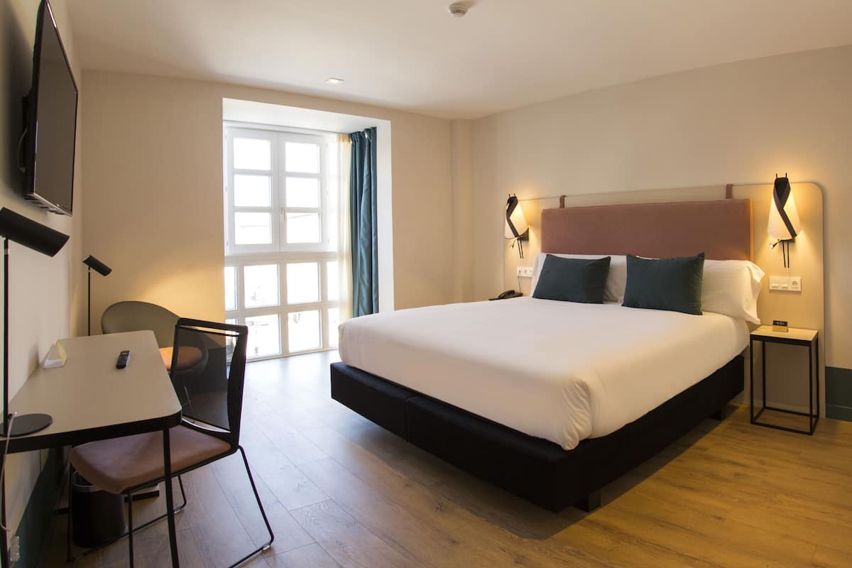 Une chambre d’hôtel avec un grand lit, un bureau avec une chaise, un téléviseur fixé au mur et une fenêtre avec des rideaux.