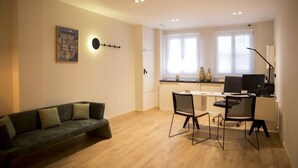 Ein moderner Wohnbereich mit einer Couch, einem Essbereich mit Stühlen, einer Küchenzeile mit Arbeitsplatte und einem Computer sowie einem großen Fenster mit Jalousien.