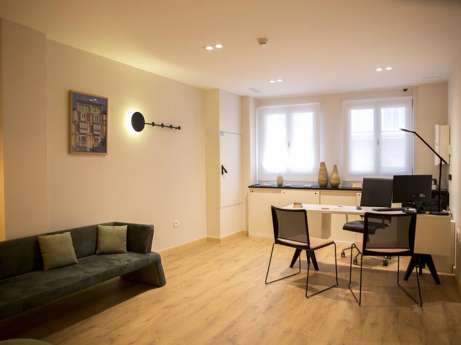A modern living space with a sofa, a dining area with chairs, a kitchen area with a counter and a computer, and a large window with blinds.
