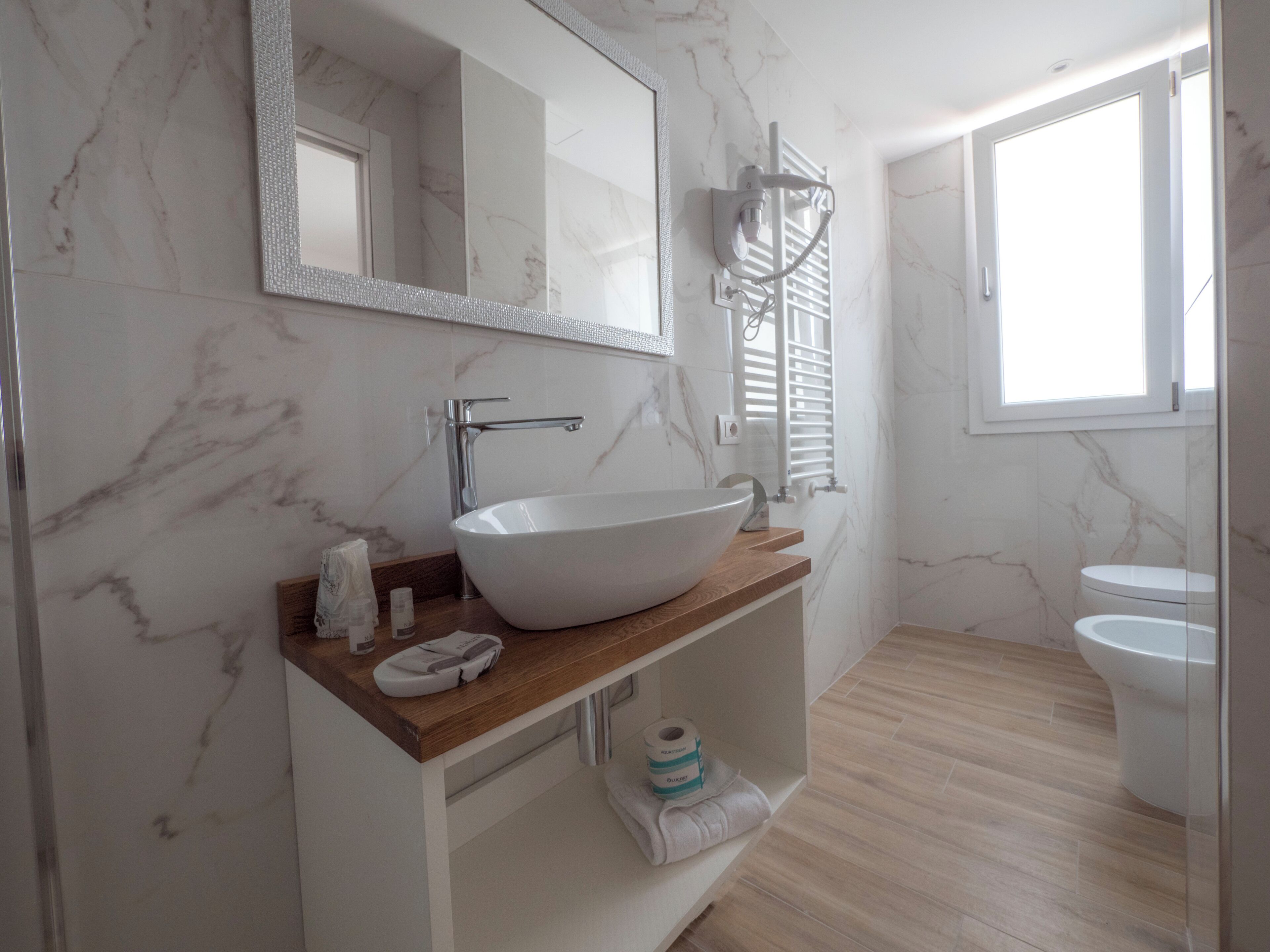 A bathroom with a marble wall, a large mirror, a vessel sink, a towel rack, and a toilet.