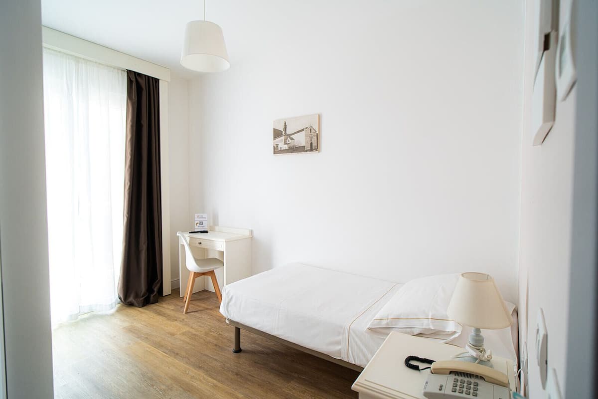 A hotel room with a bed, a desk, a chair, a lamp, a telephone, and a picture on the wall.