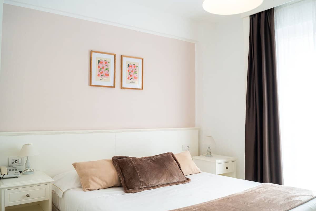A hotel room with a bed, two bedside tables, a lamp, a phone, and two framed pictures on the wall.