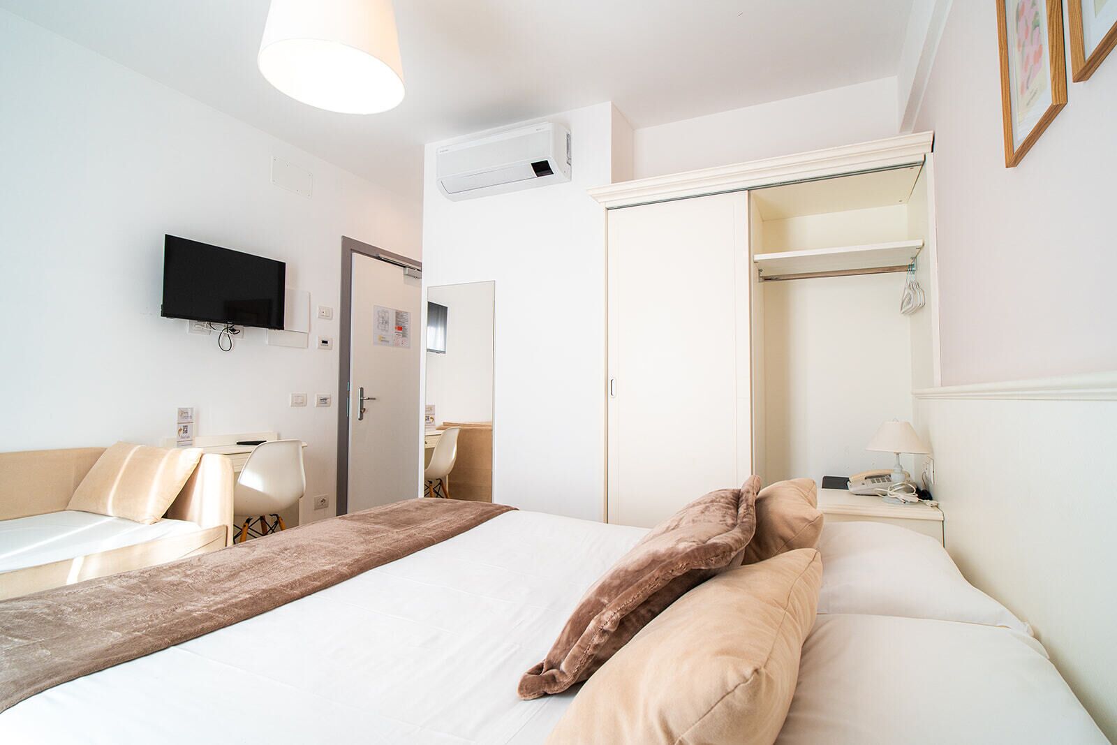 A hotel room with a bed, a desk, a chair, a wardrobe, a television, and a bedside table with a lamp.