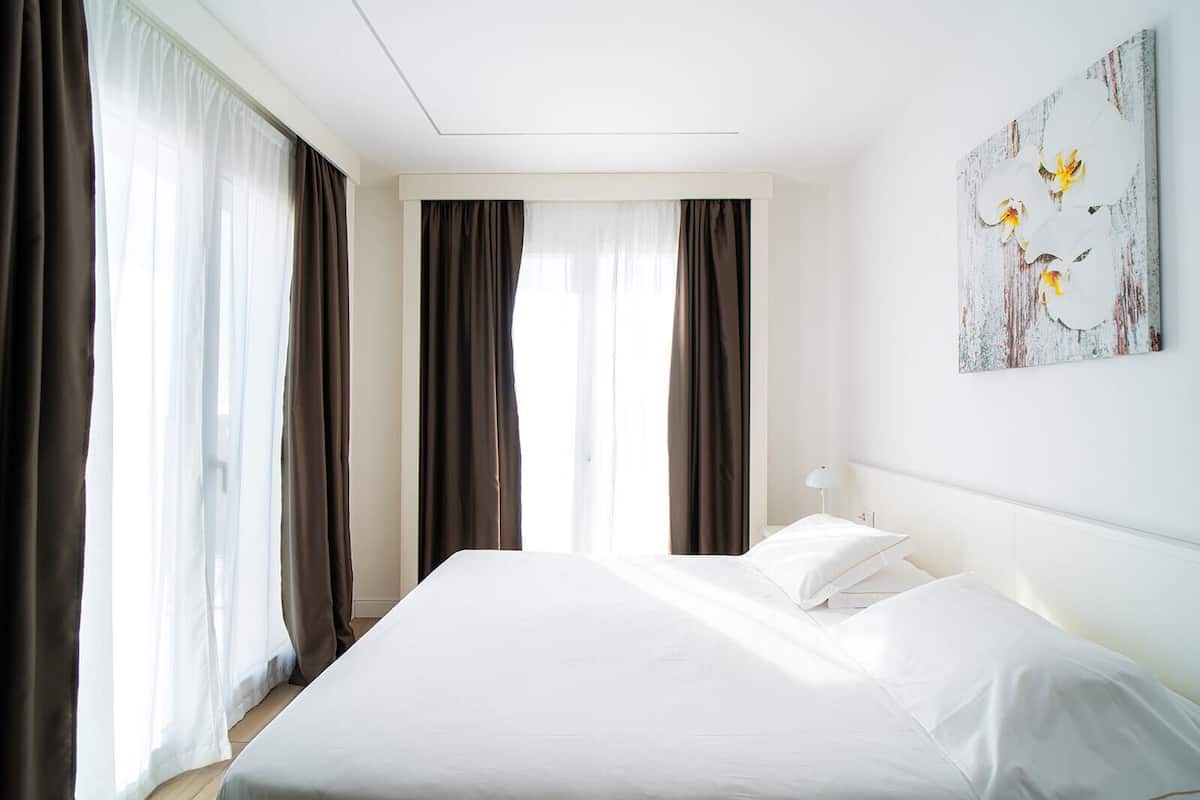 A hotel room with a large bed, two windows with curtains, a bedside table, and a wall-mounted artwork.