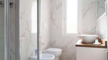 A bathroom with a glass shower enclosure, a toilet, a bidet, a sink, and a window.